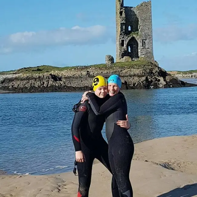 Two women in wetsuits and swim caps embracing on a beach, smiling at the camera, with an old stone tower on a small island in the background under a clear sky.