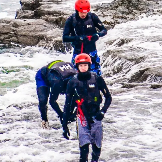 Three people in wet suits and helmets walk through rocky coastal waters, appearing focused and equipped with safety gear for a water-based activity.