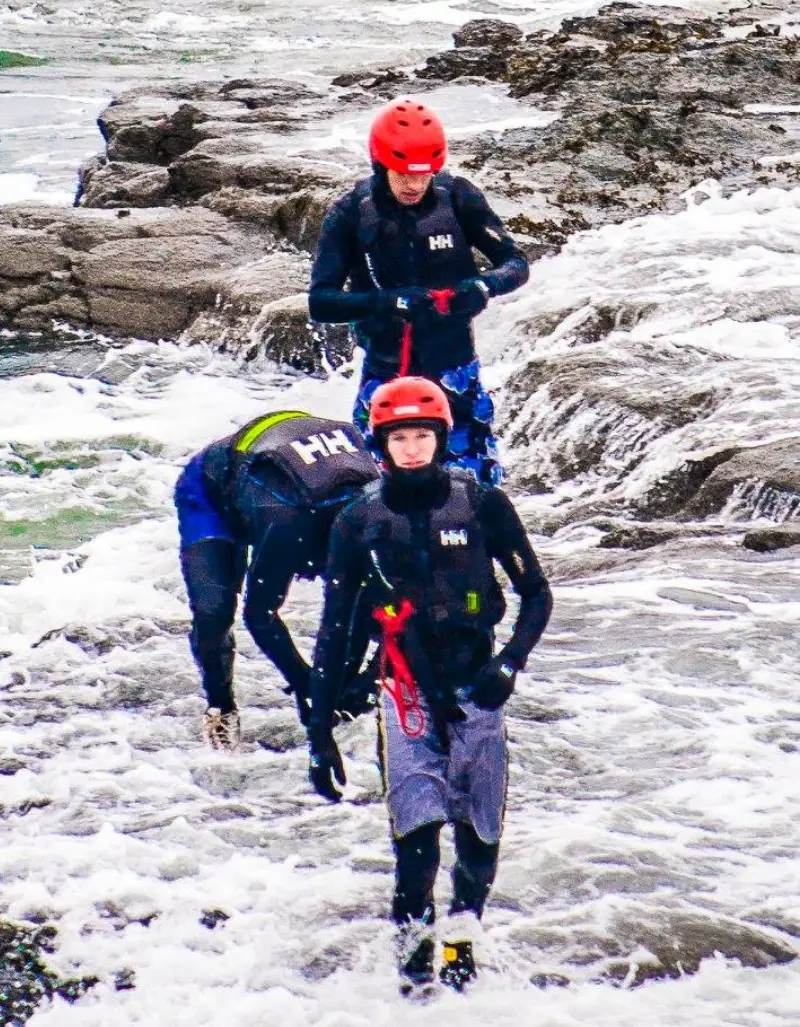 Three people in wet suits and helmets walk through rocky coastal waters, appearing focused and equipped with safety gear for a water-based activity.