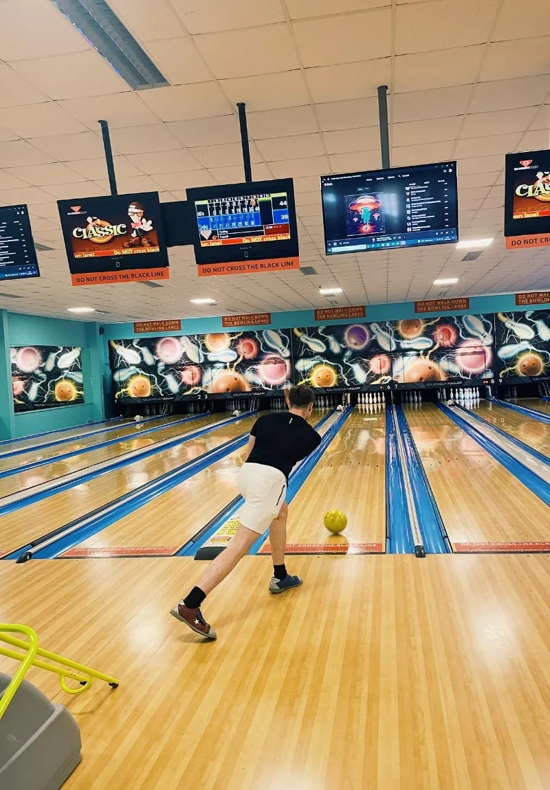 A person in a bowling alley about to release a yellow bowling ball down the lane. Colorful floral designs adorn the walls, and multiple lanes are visible with digital scoreboards above each.