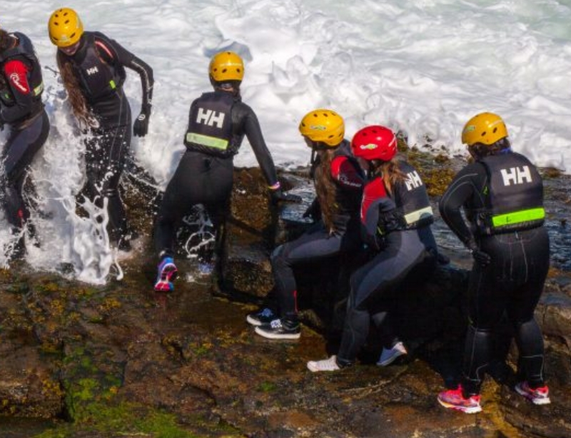 A group of people wearing wetsuits and helmets cautiously descend rocky terrain into foamy ocean waves.