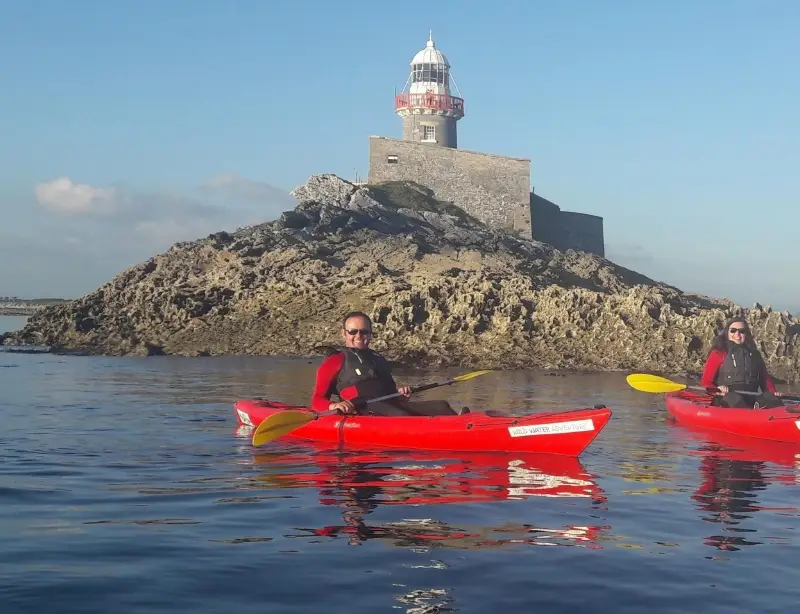 Two people kayaking near a rocky island with a lighthouse on a sunny day, calm blue water reflecting light.