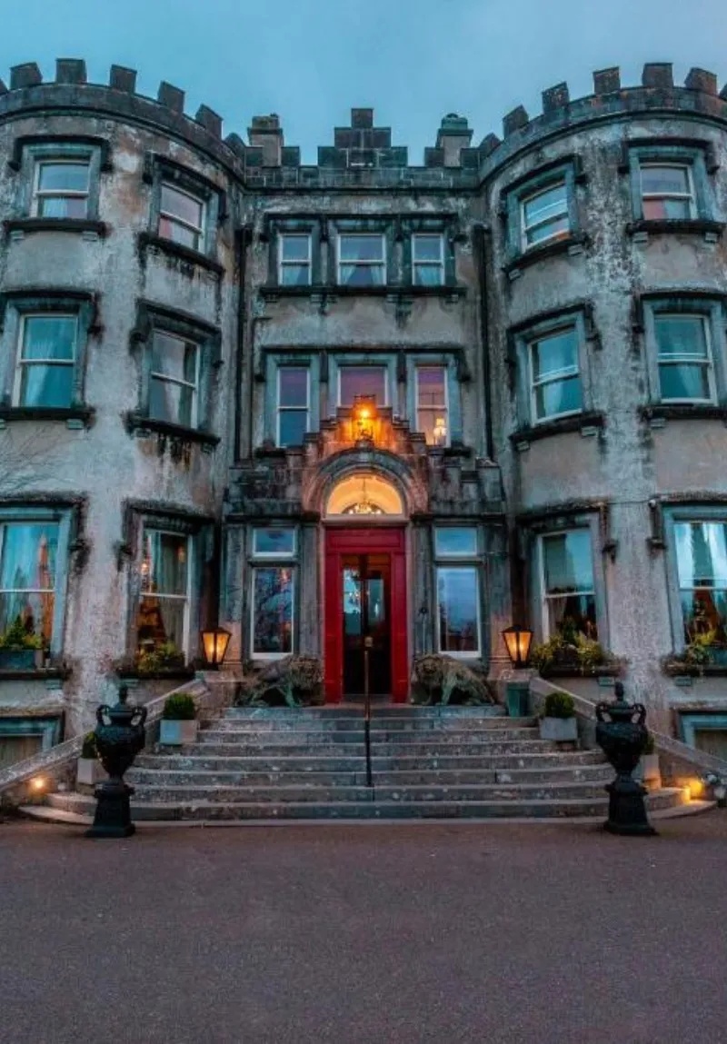 The facade of a grand, historic castle at dusk, featuring an ornate entrance with a red door, lit by warm lanterns, and multiple windows under a twilight sky.