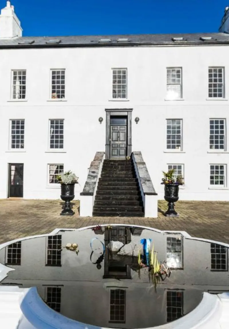 A white building with a central door flanked by windows reflected in a glossy, curved surface below, highlighting a distorted mirrored image that includes people and bicycles.