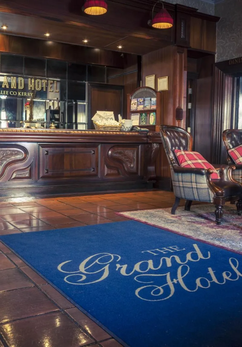 Elegant hotel lobby featuring a polished wooden reception desk with "grand hotel" inscribed, plush armchairs, and a blue carpet with the hotel's name. a cozy, welcoming ambiance is highlighted by soft lighting.