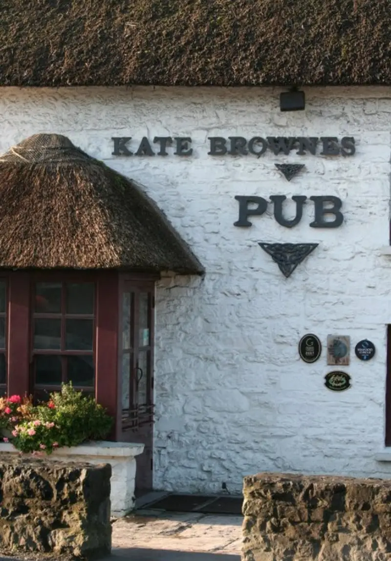 The entrance of "Kate Browne's Pub" with a thatched roof and white walls, featuring a red-framed window and decorative plaques below the bold signage.