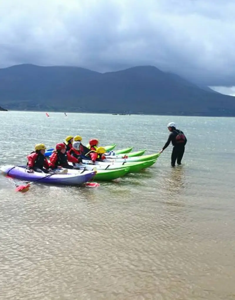 A group of people in yellow life jackets receiving kayaking instructions from a guide on a shallow, clear water beach, with mountains in the background under a cloudy sky.