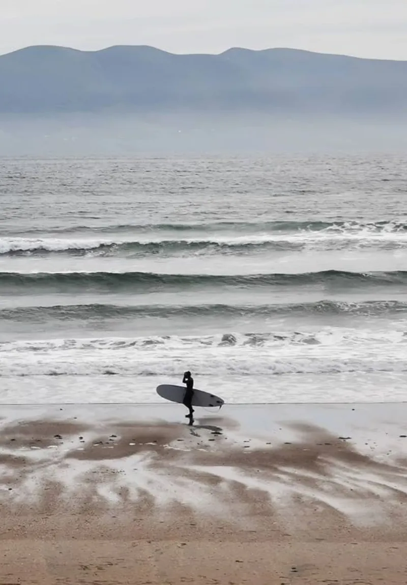 A surfer, silhouetted against the ocean waves, walks along a sandy beach with a surfboard under their arm. Misty mountains loom in the background under an overcast sky.