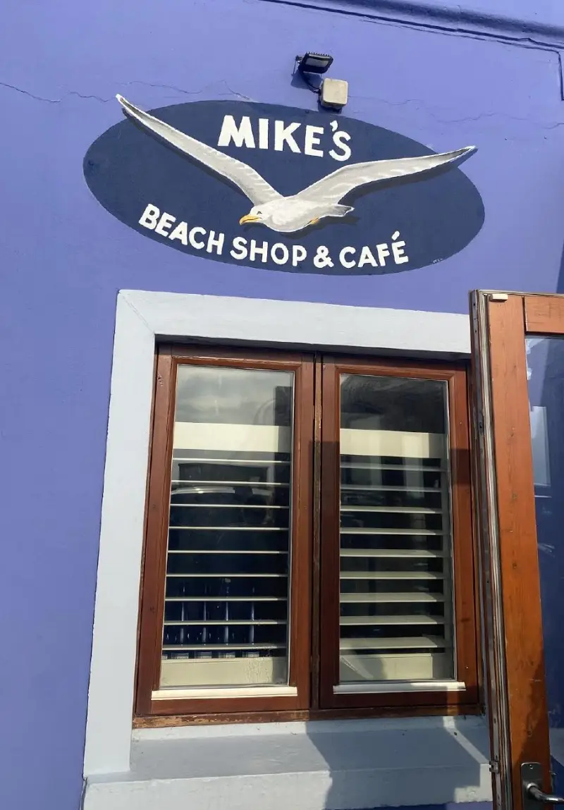 The image shows a vibrant blue wall with "MIKE'S BEACH SHOP & CAFÉ" painted in white letters, framed by a white seagull graphic. Below the sign, there's a window with white shutters and a brown door on the side.