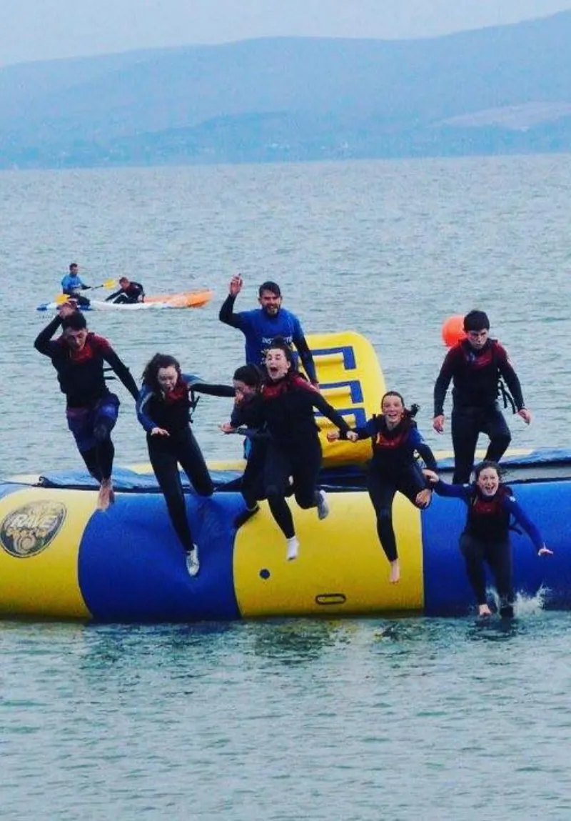 A group of people in wetsuits joyously jumping into a lake from a large yellow inflatable water trampoline, with hills visible in the background.