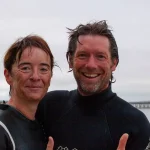 Two people in wetsuits giving thumbs up, smiling at the camera with a cloudy sky and distant pier in the background. both have wet hair, suggesting they've been in water.