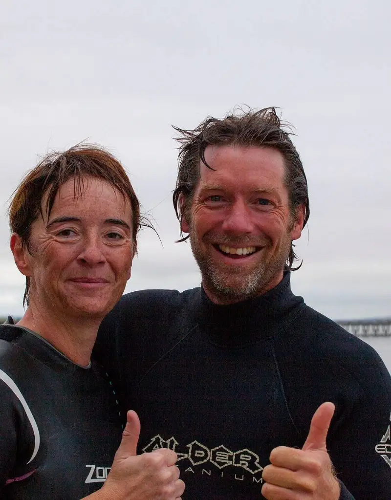 Two people in wetsuits giving thumbs up, smiling at the camera with a cloudy sky and distant pier in the background. both have wet hair, suggesting they've been in water.