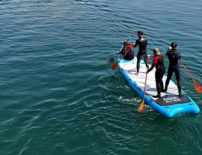 Three people in wetsuits are standing on a large blue paddleboard, paddling together on a clear, calm body of water.