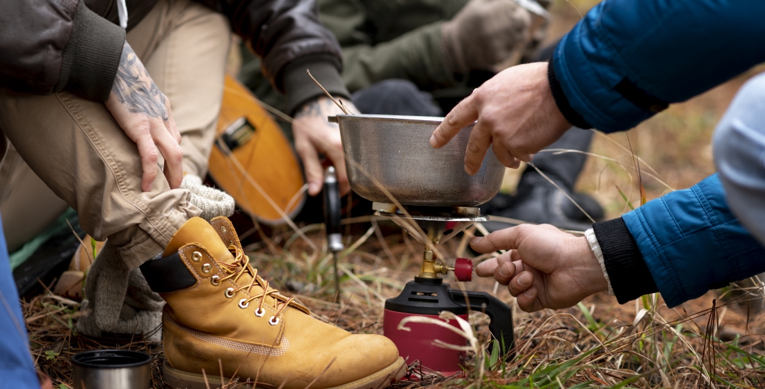 People sitting on the ground outdoors, surrounded by grass and leaves, preparing a meal over a small portable stove. One person is adjusting the burner, while another holds the pot. A pair of yellow hiking boots and a backpack are visible in the background.