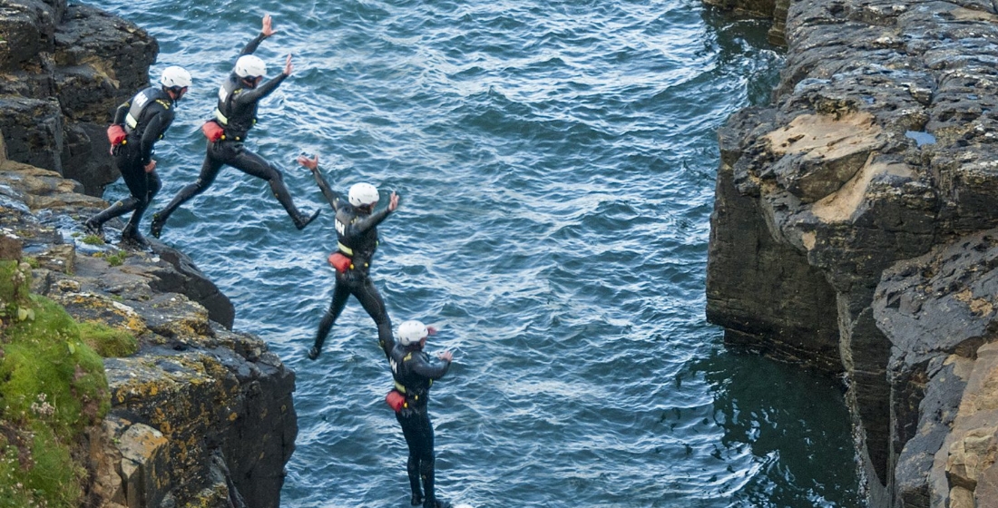 Four people wearing helmets and wetsuits are cliff jumping into the sea from a rocky ledge.