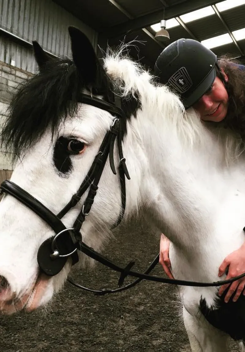 A person in a cap hugs a white and black horse wearing a bridle inside a barn. The horse peeks curiously at the camera.