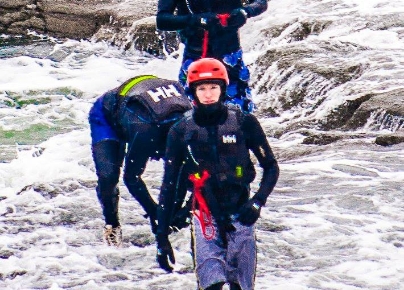 Three individuals are engaged in an adventure sport, navigating through rocky and watery terrain. The person in the foreground wears a red helmet, black wetsuit, and gray shorts, carrying safety equipment. The group appears focused and determined amidst the flowing water.