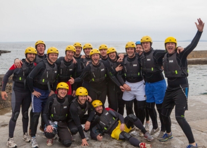 A group of people wearing yellow helmets, black wetsuits, and life vests poses together on a concrete surface near the ocean. Some are smiling and making enthusiastic gestures, indicating they might be about to participate in a water activity or have just finished one.