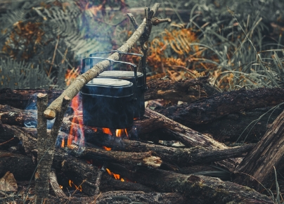 Two black camping pots are hanging over an open fire on a stick, with orange flames and smoke visible among the logs. The background features a forest floor with dry grass and ferns, suggesting a wilderness or outdoor cooking scene.