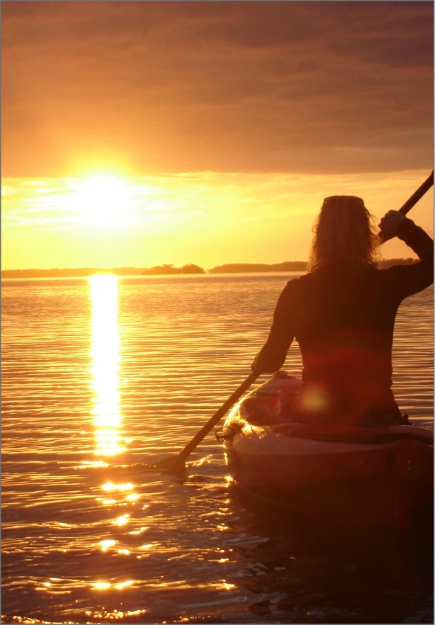 A person paddles a kayak on calm waters during a golden sunset. The sun casts a warm glow across the sky and reflects on the water, creating a serene and peaceful atmosphere. The silhouette of the kayaker is emphasized against the vibrant backdrop.