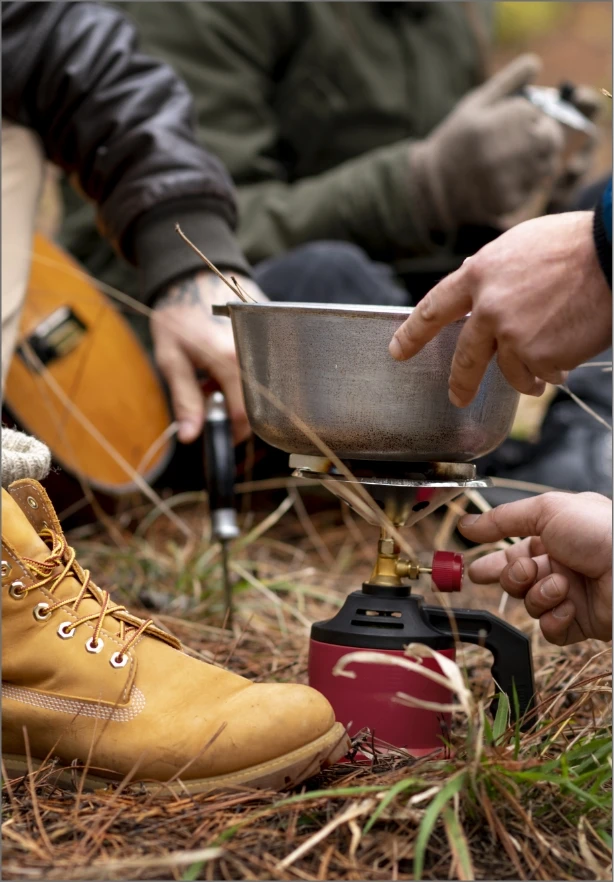 Three people, partly visible, are sitting outdoors as one adjusts a camping stove with a metal pot on top. One person is wearing a brown glove, and another wears light brown boots. The scene is set on pine needles, suggesting a forested area.