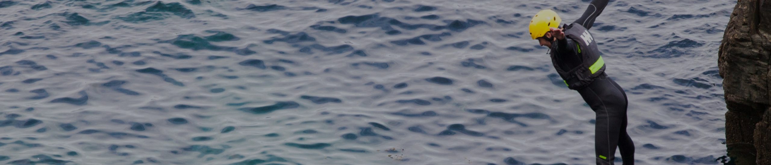 A person wearing a wetsuit and a yellow helmet is jumping off a rocky ledge into the sea. The water below appears calm with gentle waves.