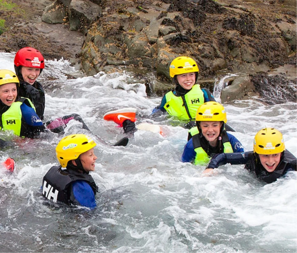 A group of five children wearing helmets and life jackets are smiling and laughing while playing in a shallow, rocky river with flowing water. They appear to be enjoying an outdoor water adventure or rafting activity.