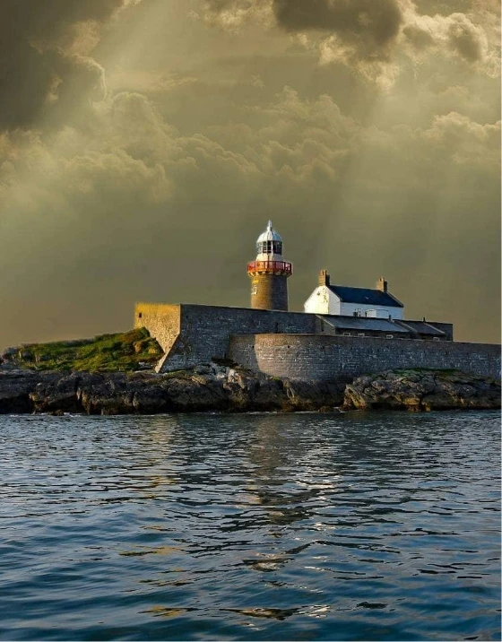 A stone lighthouse stands on a rocky shoreline, surrounded by a stone fortress, as sunlight filters through dramatic, cloud-filled skies. The calm sea in the foreground reflects the warm, golden light from the sky.