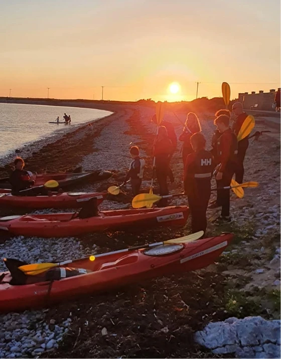 A group of people wearing life jackets stand beside red kayaks on a rocky shore at sunset. The sun is low in the sky, casting an orange glow over the scene. Some people are holding yellow paddles, and there are several kayaks on the ground and in the water.