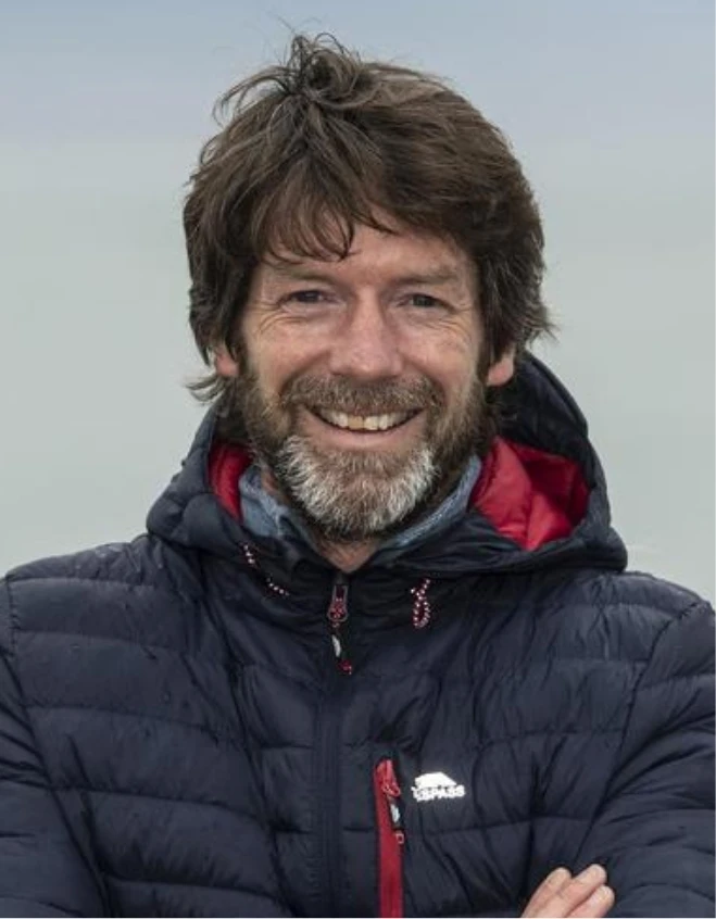 A man with dark hair and a beard smiles at the camera. He is wearing a dark puffer jacket with a hood. The background is an overcast sky.