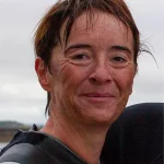 Close-up of a person with short brown hair, smiling, and wearing a wetsuit. The background is blurred, with a cloudy sky and faint outlines of a landscape. The person's face shows signs of having been wet, suggesting involvement in a water activity.