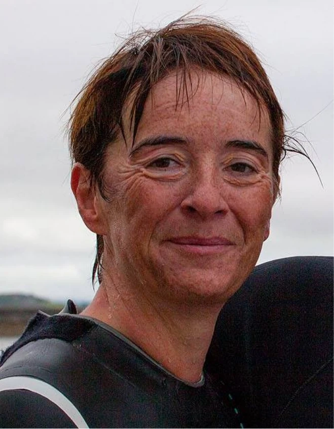 Close-up of a person with short brown hair, smiling, and wearing a wetsuit. The background is blurred, with a cloudy sky and faint outlines of a landscape. The person's face shows signs of having been wet, suggesting involvement in a water activity.