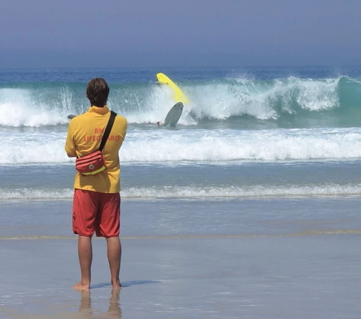 A lifeguard wearing a yellow shirt and red shorts stands on the beach, watching as a person wipes out in the ocean waves on a yellow surfboard. The surfboard is flipped, and water splashes around the surfer. The sky is clear and the ocean is blue.