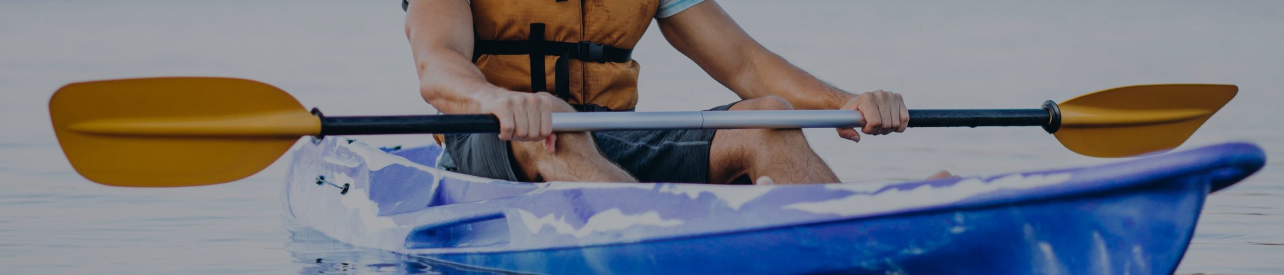 A person wearing a life jacket paddles a blue kayak on calm water. The image focuses on the hands holding the paddle and the upper half of the kayak.