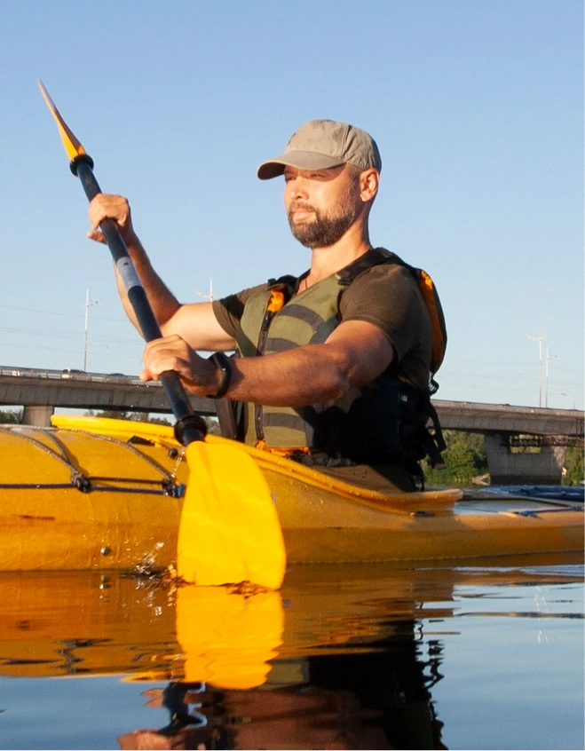 A man paddles a yellow kayak on calm water under a clear blue sky. He wears a green shirt, a gray cap, and a life vest. A bridge with power lines is visible in the background.