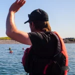 Person in a black cap and red life jacket raises an arm while standing in a boat on the water. Others in the distance are swimming near a rocky shoreline under a clear blue sky.