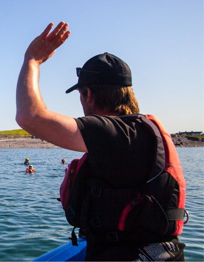 Person in a black cap and red life jacket raises an arm while standing in a boat on the water. Others in the distance are swimming near a rocky shoreline under a clear blue sky.