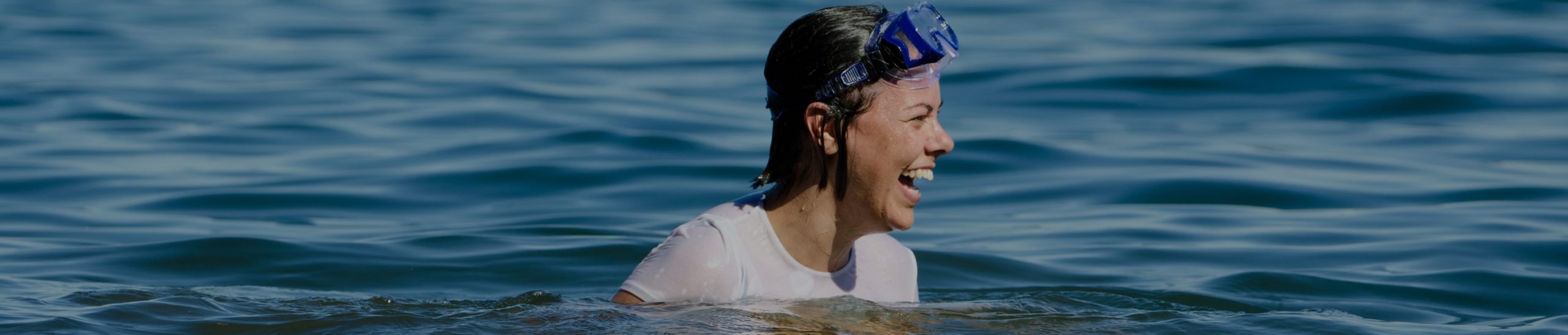 Person with short hair wearing a snorkel mask and white shirt, smiling and wading in calm water. The background shows an expanse of blue water under a clear sky.