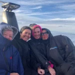 Four people dressed in warm Dryrobe jackets sit close together, smiling at the camera on a boat ride. The sky above them is partly cloudy, and they appear to be enjoying the cool weather on the water.