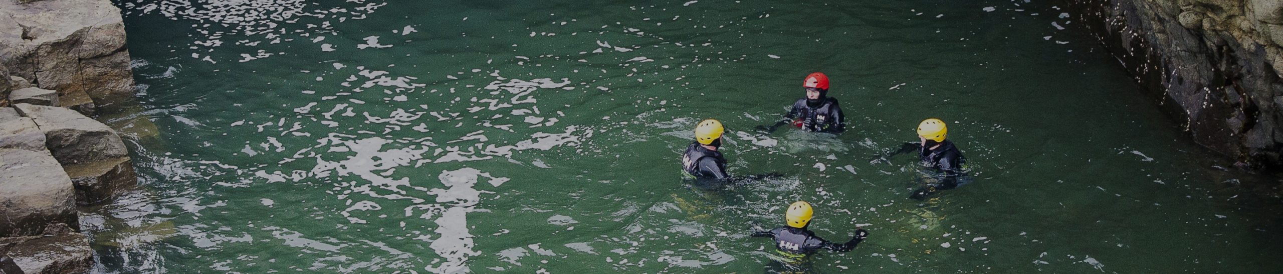 Four individuals wearing wetsuits and helmets are swimming in a rocky and clear green body of water. Three have yellow helmets and one has a red helmet. The water surface is gently rippled, and the surroundings indicate a natural, rugged environment.