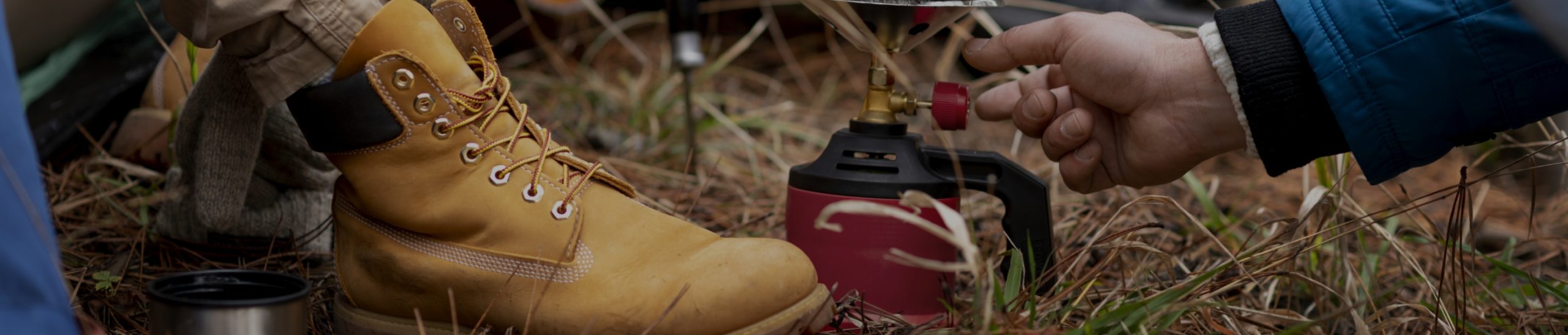 Close-up of a person wearing tan hiking boots sitting on grass near another person who is using a portable camp stove. The scene includes various camping gear, and the background shows more grass and dirt, suggesting an outdoor setting.