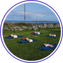 Several CPR mannequins lie on blue mats on green grass in an open field, with a stone wall, electric poles, and the ocean visible in the background. The sky is partly cloudy.