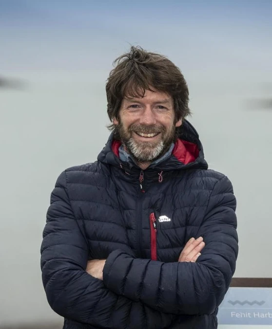 A man with a beard and long hair, wearing a dark quilted jacket, stands outside with his arms crossed. He is smiling and the background appears to be a cloudy sky and misty landscape.