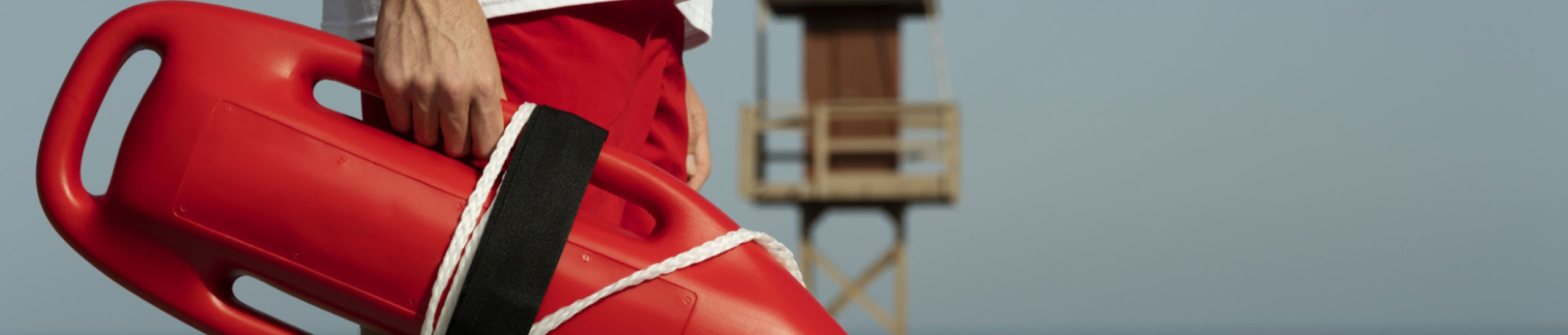 A lifeguard in red shorts holds a red flotation device while standing on a beach. A wooden lifeguard tower is visible in the background against a clear blue sky.