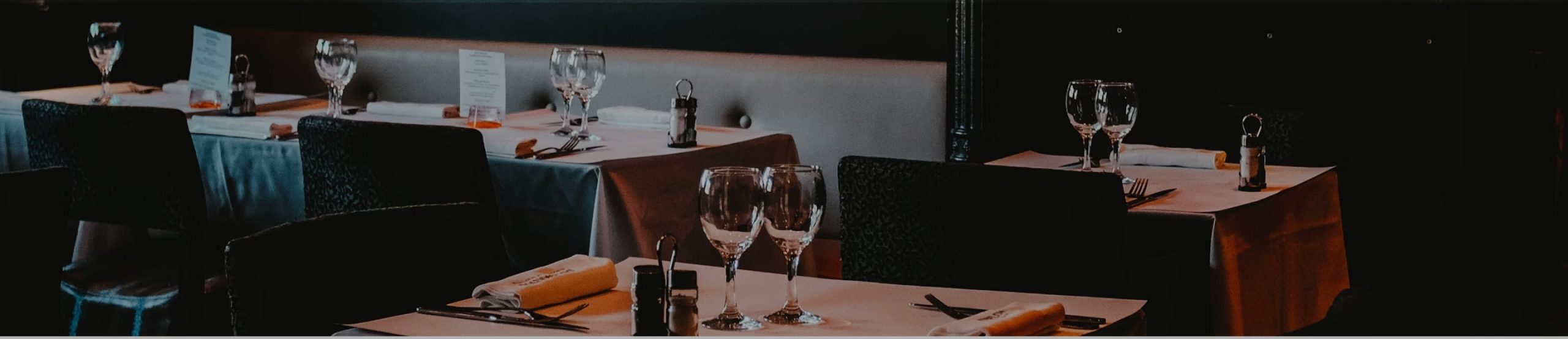 A dimly lit restaurant with neatly set tables draped in white tablecloths. The tables feature wine glasses, napkin-covered plates with silverware, and small decorative items. The overall ambiance is elegant and inviting.