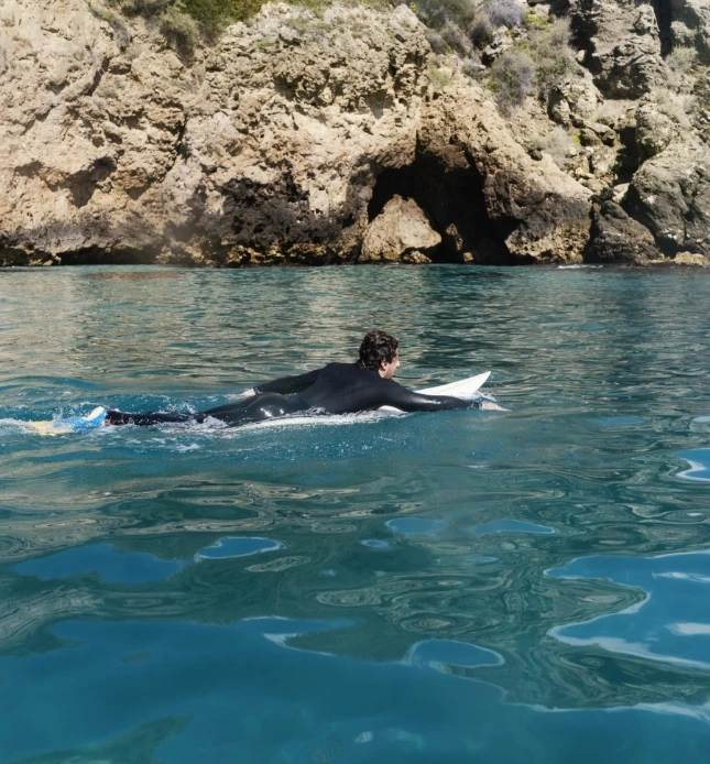 A person wearing a black wetsuit is lying on a surfboard and paddling on clear blue water toward a rocky shore. The rocky terrain features large, rugged cliffs with patches of vegetation. The scene is set on a bright, sunny day.