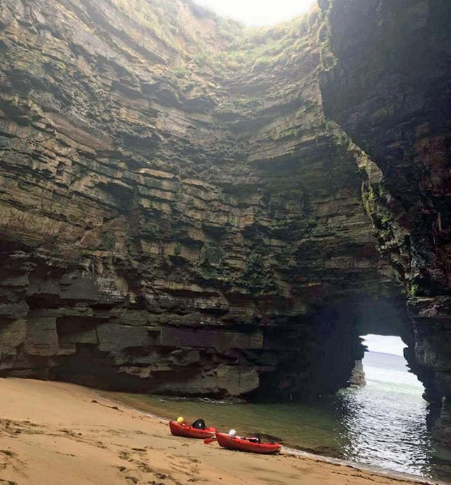 A picturesque scene of a hidden cave with towering rock walls, sandy shoreline, and an opening that allows a glimpse of the sea. Two red kayaks are resting on the sand, enhancing the adventurous feel of the secluded location.