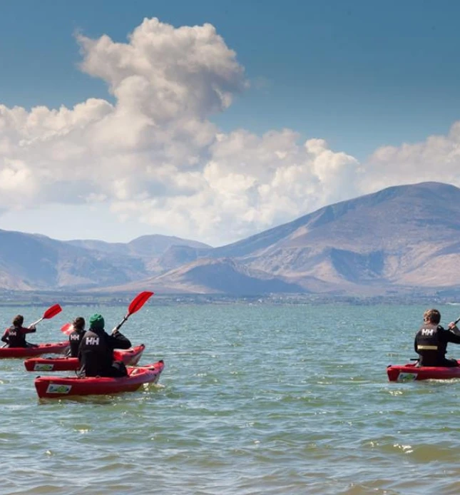 Four people kayaking on a large lake with mountainous terrain in the background. The kayakers are wearing black shirts, and the kayaks are red. The sky is mostly clear with a few clouds.