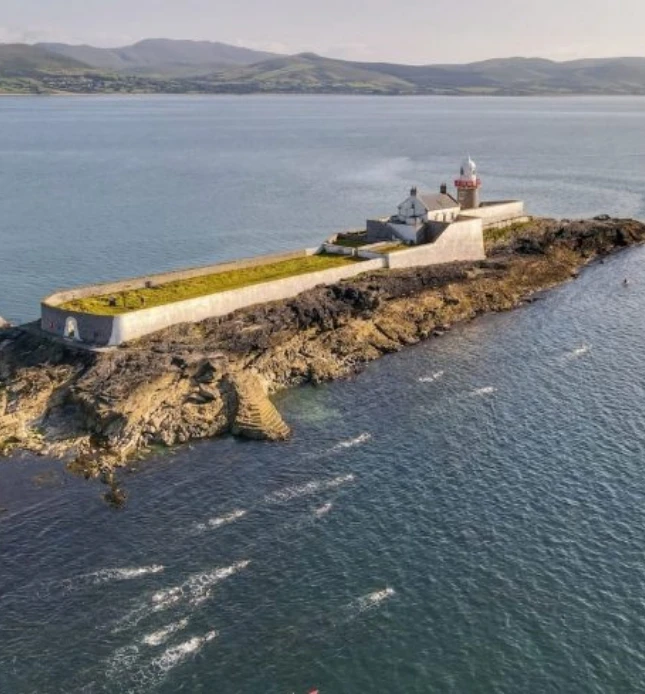 An aerial view of a small island with a historic fort and lighthouse surrounded by calm ocean waters. The island features rocky shores and stone steps leading up to the fort. The background shows distant, rolling hills under a clear sky.