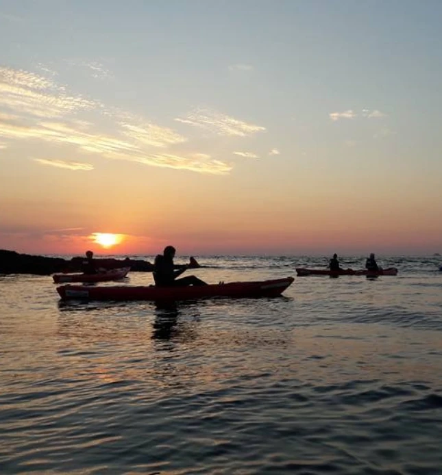 Three people are kayaking on calm waters during sunset. The sky is a blend of orange, pink, and purple hues, and the sun is partially set on the horizon, casting reflections on the water. Silhouetted kayakers are seen paddling leisurely.